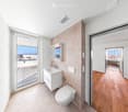 Modern bathroom with white tiles, a floating sink, toilet, and mirrored cabinet. Large window offers a view of the balcony and outdoor area.