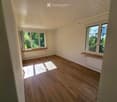 Empty room with wooden flooring, white walls, and large windows with wooden sills. Natural light streams in, casting shadows on the floor.