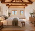 Cozy bedroom with exposed wooden beams, large windows, a bed with pillows and a blanket, nightstands, a bench, and a woven rug.