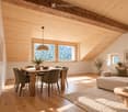 Bright, open living and dining area with wooden beams, large windows offering a view, a sofa, coffee table, dining table with chairs, pendant light, and decorative vases.