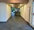 Empty hallway with tiled flooring, white walls, wooden ceiling, and recessed lighting. Features a reception desk and a glass door leading outside.