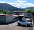 A modern building complex with a garage, parking area, and surrounding greenery, set against a scenic mountain backdrop.