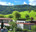 A cluster of buildings in a rural setting with green hills and trees in the background. The buildings include a mix of residential and commercial structures, with visible parking spaces and a van.