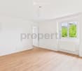 Empty room with light wooden flooring, white walls, and a large window with white frames. A small adjacent bathroom is visible with a toilet and sink.