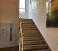 Modern staircase with beige stone steps, wooden handrail, and a wall-mounted light fixture. The hallway features a textured wall and a glass panel on one side.