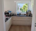 Modern kitchen with light-colored cabinets, wooden flooring, and various appliances including a microwave, toaster, coffee maker, and knife block. A window offers a view of greenery outside.