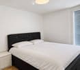 Modern bedroom with a double bed, white bedding, a black headboard, a nightstand, and a large window with blinds.