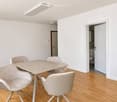 Dining area with a table and four chairs, light-colored walls, wooden flooring, and a cabinet on the left side.