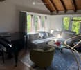 A modern living room with a grey sectional sofa, a black grand piano, a glass coffee table, a green armchair, and a floor lamp. The room features large windows with curtains and a patterned rug.