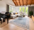 Bright living room with wooden floors, large windows, a grand piano, a sectional sofa, a coffee table, a TV, and decorative plants.