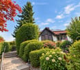 A two-story house with a red-tiled roof and white walls, surrounded by a well-maintained garden with various flowering plants and shrubs. The entrance features a paved pathway leading to a small staircase.