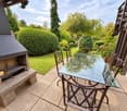 Outdoor patio with a glass table and metal chairs, surrounded by a well-maintained garden with trees and shrubs.