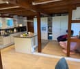 Open-plan kitchen and living area with white cabinets, a central island, and modern appliances. Wooden beams on the ceiling and a pink chair near the island.