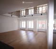 Large empty room with wooden floor, white walls, three windows with radiators, black railing on raised ground floor