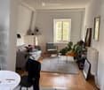 A cozy living room with a grey sofa, wooden coffee table, armchair, and several potted plants. The room features wooden flooring, a large window, and a small dining area with a white table and black chair.