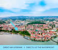 Aerial view of a town with a waterfront, featuring numerous buildings, a marina, and surrounding greenery.