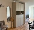 A modern, minimalist living space with light wooden flooring, a grey upholstered chair, a sleek cabinet, a round mirror, a decorative vase with dried plants, and a small table with a screen and lamp.