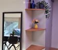A narrow hallway with light wooden flooring, white walls, and built-in wooden shelves holding decorative items such as vases, flowers, and a potted plant. A large mirror is mounted on the left wall, reflecting part of the adjacent room.