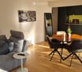 Modern living and dining area with a grey sofa, coffee table, dining table with chairs, and a potted plant. The space features wooden flooring and large windows with white curtains.