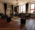 Modern living room with wooden flooring, large windows, a black leather sofa, two black leather chairs, two round gold tables, two black stools, a large floor lamp, a plant, and a shelf with books and decorative items.