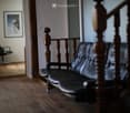 A furnished room with a black leather sofa and a black office chair, wooden flooring, and a staircase with wooden railings.