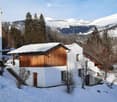 Modern white house with wooden accents, snow-covered roof, and steps leading to the entrance, surrounded by snow-covered trees and mountains.