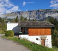 A small, modern white house with a stone roof, surrounded by lush greenery and mountains in the background.