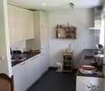 Modern kitchen with white cabinets, dark countertops, and a small wooden shelf holding decorative items. A small round table with a potted plant is visible, along with a wooden chair and a small wooden stool.