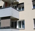 Balconies with grey wooden paneling and white railings, attached to a beige building with multiple windows and shutters.