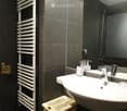 Modern bathroom with dark tiled walls, a white towel rack, a sink with a modern faucet, and a large mirror above it. Soap dispensers are placed on the sink counter.