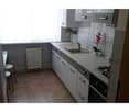 Modern kitchen with white cabinets, a sink, stove, and a small flower pot on the counter. There is a radiator under the window and a stool near the entrance.