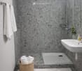 A bathroom featuring a modern, stylish design with tiled floors and walls. A shower stall with a glass door and silver shower head is present. There is a white sink with a faucet and a mirror on the wall. Two white towels are on a towel rack and a wicker