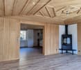 Bright, open room with wooden walls and ceiling, featuring a modern fireplace and a view of a kitchen area through an open doorway.