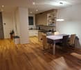 Modern open-plan kitchen and dining area with light wood flooring, grey cabinets, and a central island. A dining table with four chairs is set with a patterned tablecloth.