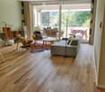 Modern living room with wooden flooring, large windows, and a balcony. Features a grey sofa, coffee table, chairs, floor lamp, and speaker.