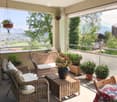 Balcony with wicker sofa, chairs, small table, and potted plants, overlooking a scenic valley with mountains.