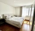 A well-lit bedroom with wooden flooring, a large bed with a headboard, nightstands with lamps, and a window with curtains.