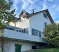 Modern two-story house with white exterior, blue shutters, and a large balcony on the first floor.