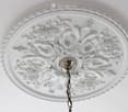 An ornate white ceiling medallion with floral and scrollwork design, featuring a central light fixture with a hanging chain.