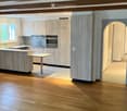 Modern kitchen with light wood cabinets, stainless steel appliances, and a central island with a sink and faucet. The space features wooden flooring and an arched doorway leading to another room.