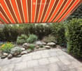 Garden area with a stone-paved patio, large rocks, flowering plants, and a retractable awning providing shade.