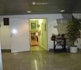 A modern hallway with a polished tile floor, white walls, and a large potted plant. A small table holds a sewing machine and a picture frame, with a doorway leading to another room.