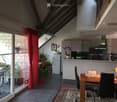 Open-plan living and dining area with a dining table, chairs, kitchen area, and potted plants. Features a large window with a red curtain and a view of a balcony with outdoor furniture.