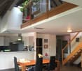 Open-plan living and dining area with a wooden dining table, black chairs, modern kitchen, staircase leading to an upper level, and decorative elements like a flower vase and picture frames.