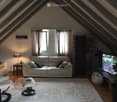 Bright living room with slanted wooden ceiling, large window with curtains, white sofa, armchair, ottoman, coffee table, lamp, TV, and potted plant.