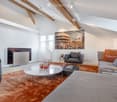 Modern living room with exposed wooden beams, large windows, a fireplace, a sofa, armchair, coffee table, and artwork on the wall.