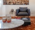 Modern living room with a large wall art depicting the Colosseum, a sleek black leather armchair, a round glass coffee table with decorative candle holders, and a small side table with a lamp and books.