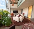 Modern living room with large windows offering a view, featuring a white sectional sofa, ottoman, armchair, coffee table, television, and decorative plants.