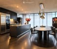 Modern kitchen with dark cabinets, stainless steel appliances, a central island, and a round dining table with chairs. Features pendant lighting and large windows with sheer curtains.