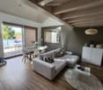Modern living room with a grey sectional sofa, coffee table, dining table with chairs, and a large window with blinds leading to a balcony.